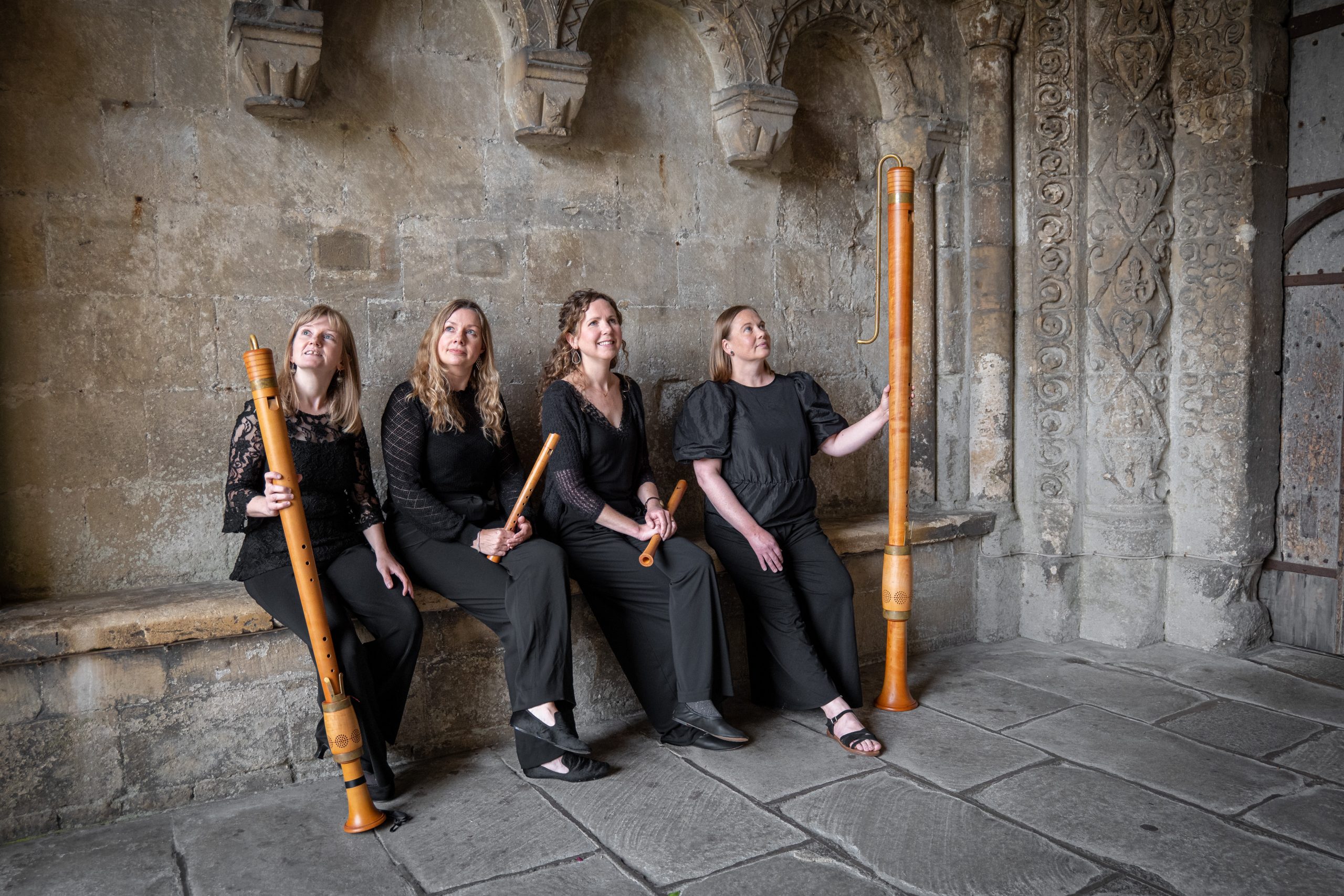 Four musicians with recorders, sitting in abbey porch with ornate stonework
